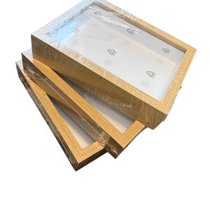 Könnte beinhalten: Drei hellfarbige Holzbilderrahmen übereinander gestapelt. Jeder Rahmen hat eine transparente Kunststoffabdeckung und eine weiße Rückseite. Die Rahmen sind rechteckig mit einem schlichten Design, geeignet für Fotos oder Kunstwerke.