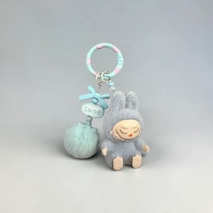 May include: A gray plush bunny keychain with a sleeping face, wearing a hood. The keychain has a light blue pom-pom, a bow, and a ring with pink and white accents. The word "cute" is on a small blue tag.