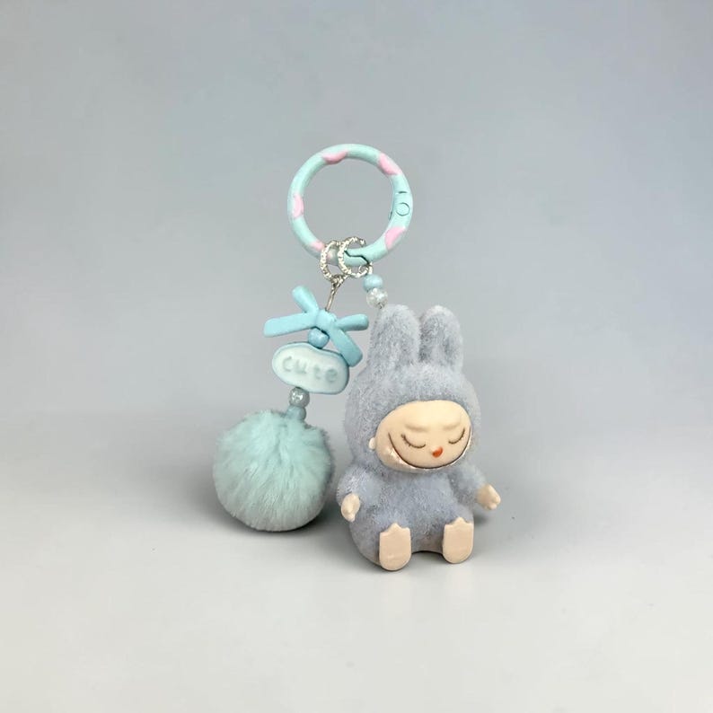 May include: A gray plush bunny keychain with a sleeping face, wearing a hood. The keychain has a light blue pom-pom, a bow, and a ring with pink and white accents. The word "cute" is on a small blue tag.