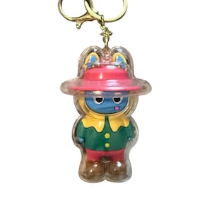 May include: A novelty keychain featuring a cartoon character in a clear plastic casing. The character has a blue face, a red hat, and is wearing a green and red outfit. The keychain has a gold-colored metal ring.