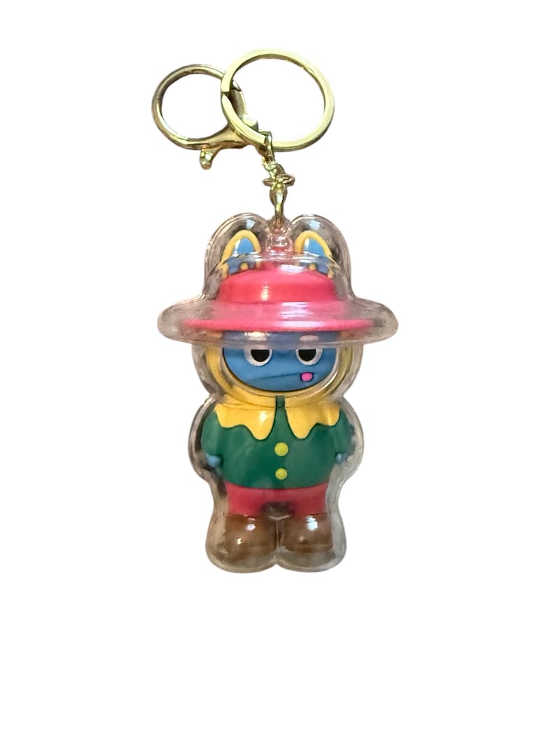 May include: A novelty keychain featuring a cartoon character in a clear plastic casing. The character has a blue face, a red hat, and is wearing a green and red outfit. The keychain has a gold-colored metal ring.
