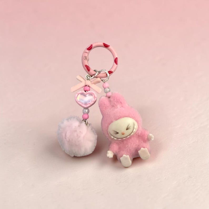 May include: A pink keychain featuring a plush bunny figure and a fluffy pom-pom. The keychain has a pink ring with red heart accents, a heart-shaped charm, and a small bow. The bunny is wearing a pink hooded outfit.