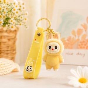 May include: A yellow keychain featuring a cartoon bunny character in a hooded outfit. The keychain has a matching yellow strap with a cartoon face and a gold-colored ring.