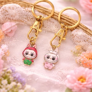 May include: Two keychains with gold-colored hardware and cartoon bunny charms. One bunny wears a red hat and green overalls, the other a pink outfit. The background features flowers and decorative elements.