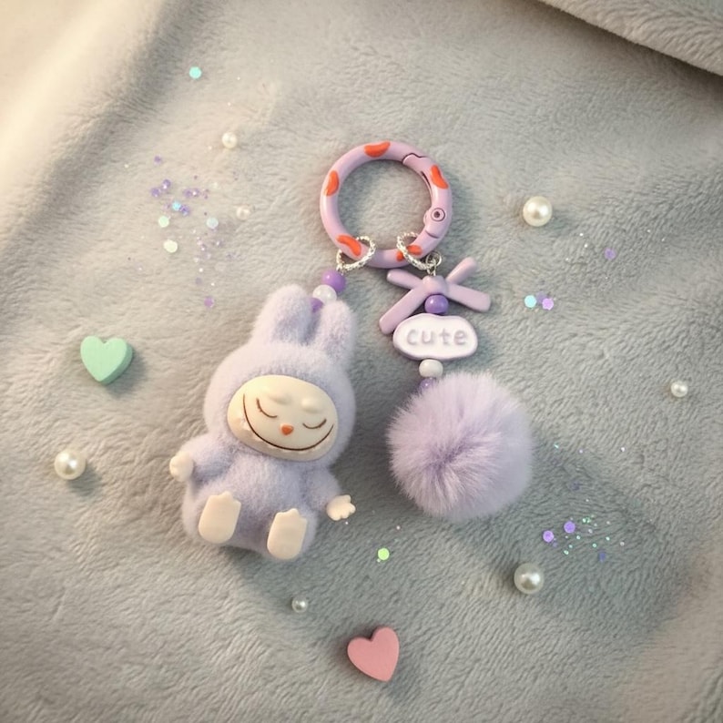 May include: A lavender bunny keychain with a smiling face, wearing a hooded outfit. The keychain has a fluffy pom-pom, a ring with orange and red accents, and the word "cute" on a small sign. Decorative pearls and hearts surround the keychain.
