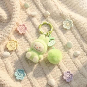 May include: A light green bunny keyring with a fluffy pom-pom and a "cute" tag. The keyring is surrounded by small, crocheted flowers in pastel shades and small white stones, all on a cream-coloured, textured surface.