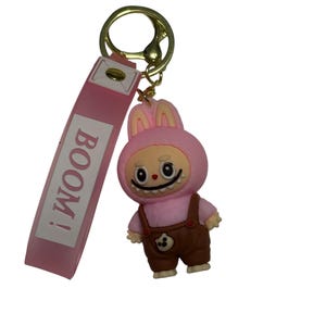 May include: A pink keychain featuring a cartoon character in a bunny costume with brown overalls. The keychain has a pink strap with the word "BOOM!" printed on it and a gold-colored metal ring.