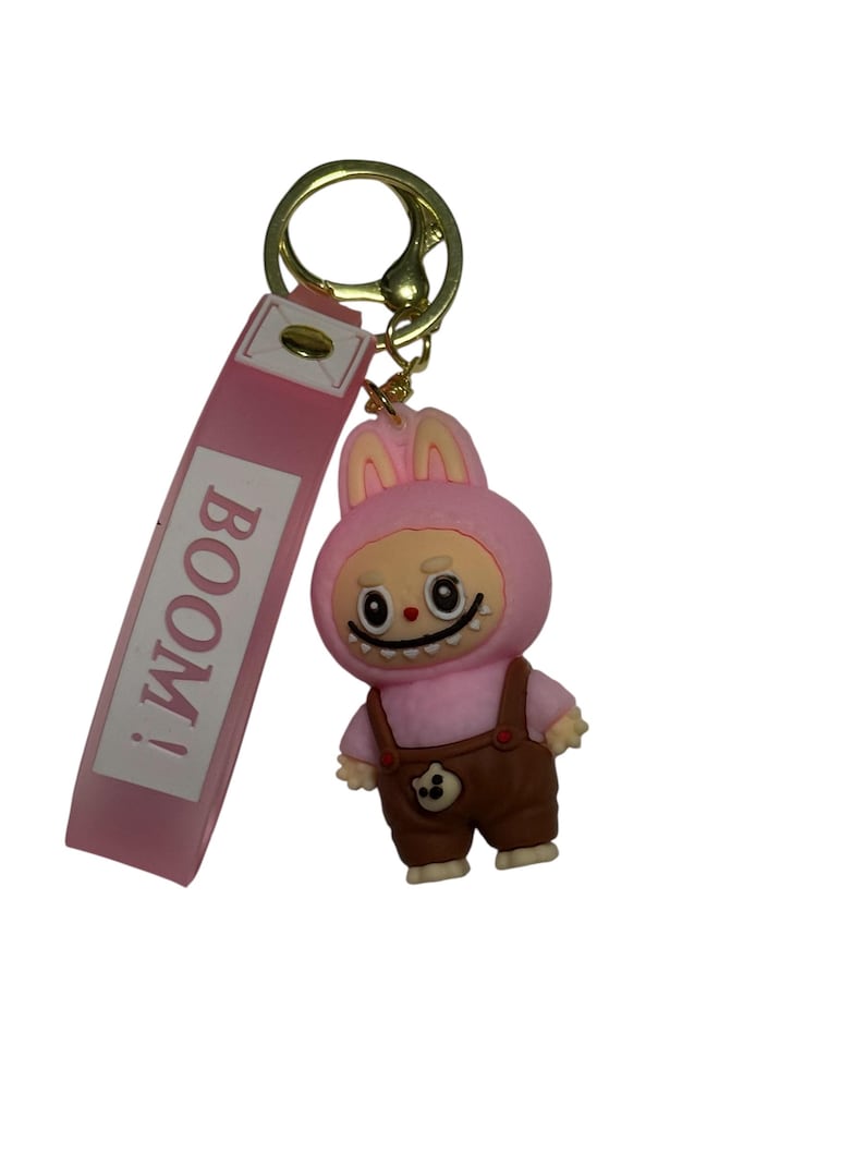 May include: A pink keychain featuring a cartoon character in a bunny costume with brown overalls. The keychain has a pink strap with the word "BOOM!" printed on it and a gold-colored metal ring.