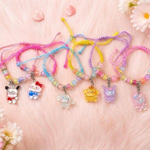 May include: Six colorful charm bracelets with adjustable cords. Each bracelet features a different cartoon character charm and iridescent beads. The bracelets are displayed on a pink, fluffy surface, with flowers and heart-shaped decorations.