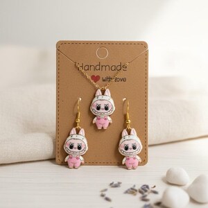 May include: A handmade jewelry set featuring a pendant necklace and matching earrings. Each piece showcases a pink bunny character with a white hood and overalls. The set is displayed on a brown card with the text "Handmade with love".