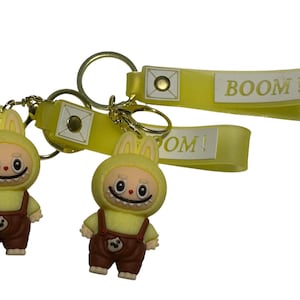 May include: Two keychains featuring yellow bunny-like figures wearing brown overalls. The keychains have gold-colored rings and yellow straps with the word "BOOM!" printed on them. The figures have a smiling face.