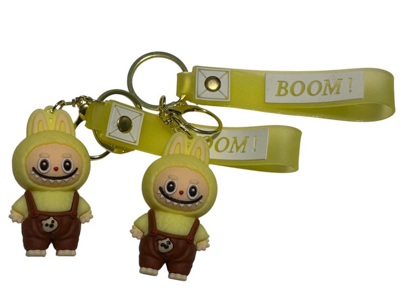 May include: Two keychains featuring yellow bunny-like figures wearing brown overalls. The keychains have gold-colored rings and yellow straps with the word "BOOM!" printed on them. The figures have a smiling face.