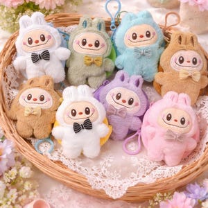 May include: A collection of plush bunny-shaped keychains in a woven basket. The keychains are in various pastel colors, including white, green, blue, brown, purple, and pink. Each keychain features a smiling face, a bow tie, and a small loop for attaching keys.