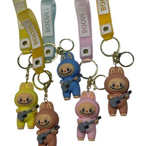 May include: A collection of keychains featuring cartoon characters in bunny suits, each holding a guitar. The keychains have colorful straps with the word "BOOM!" printed on them. The keychains are in yellow, blue, pink, and brown.