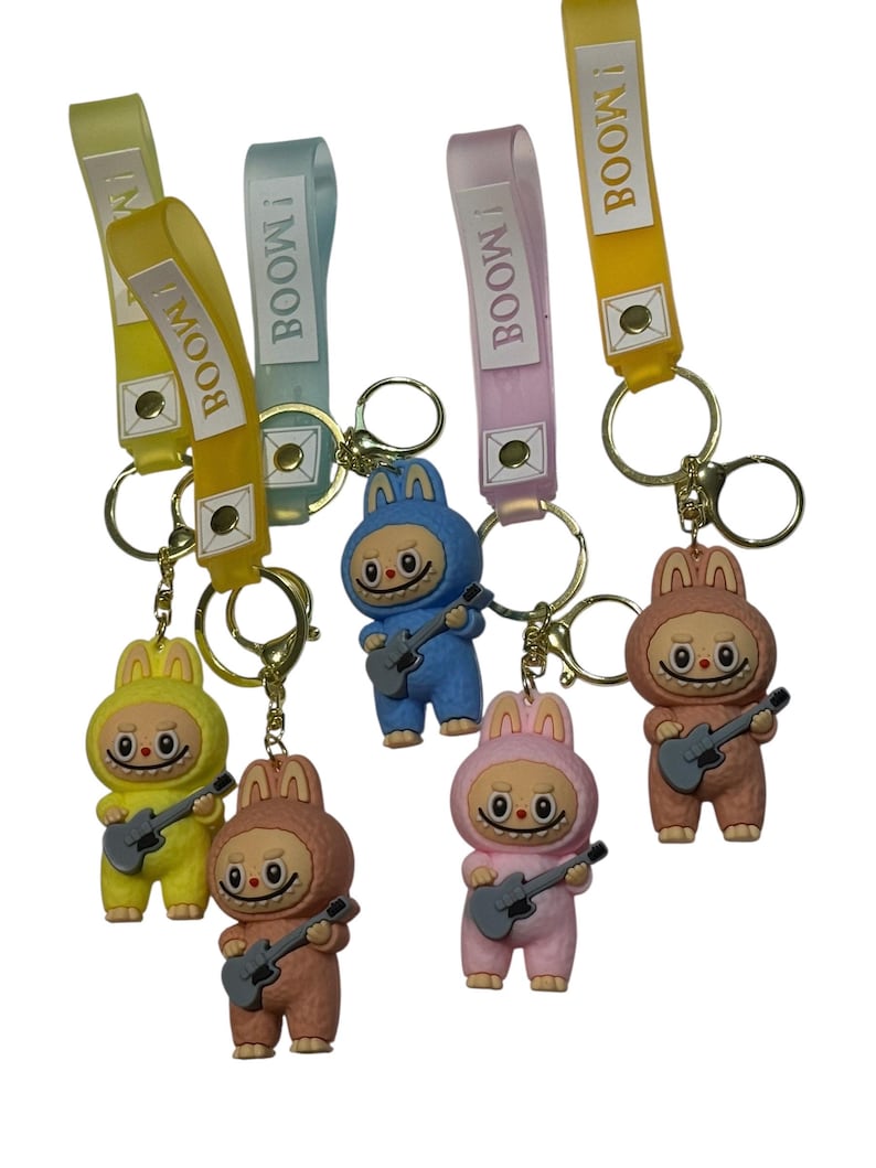 May include: A collection of keychains featuring cartoon characters in bunny suits, each holding a guitar. The keychains have colorful straps with the word "BOOM!" printed on them. The keychains are in yellow, blue, pink, and brown.