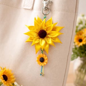 May include: A yellow sunflower keychain with a dark maroon center and a smaller sunflower charm. The keychain is attached to a cream-colored tote bag with a silver clasp. The sunflower is made of fabric.