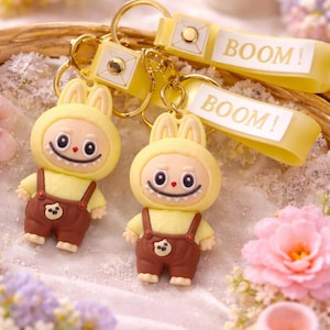 May include: Two yellow and brown bunny keychains with the word "BOOM!" printed on the yellow strap. The keychains feature a cartoon bunny wearing a yellow hoodie and brown overalls. The background includes flowers.