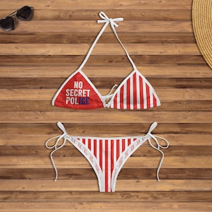 Puede incluir: Un conjunto de bikini a rayas rojas y blancas con el texto "NO SECRET POLICE" en la parte superior. La parte superior del bikini es de estilo triangular con lazos blancos, y la parte inferior es de corte clásico. Gafas de sol y sombrero de paja en el fondo.