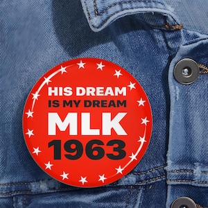 Puede incluir: Un botón redondo rojo con texto y estrellas blancas. El texto dice "HIS DREAM IS MY DREAM" encima de "MLK 1963". El botón está sobre un fondo de mezclilla azul.