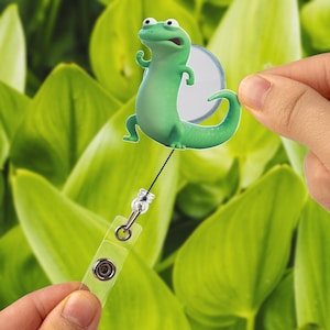 Puede incluir: Un carrete de insignia de lagarto de dibujos animados verde con un soporte de plástico transparente y un cordón retráctil. El lagarto tiene un círculo blanco en la espalda. El fondo es un desenfoque de hojas verdes.