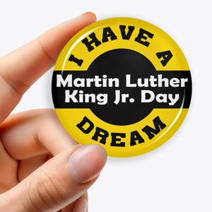 以下が含まれることがあります： 「I HAVE A DREAM」の黒い文字と「Martin Luther King Jr. Day」の白い文字が入った、丸い黄色のボタン。ボタンは手で持たれています。