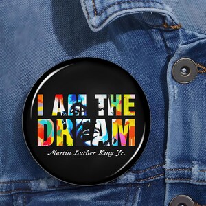 以下が含まれることがあります： カラフルなブロックレターで「I AM THE DREAM」と書かれた丸い黒いボタン。ボタンには、マーティン・ルーサー・キング・ジュニアの肖像画と彼の名前も表示されています。