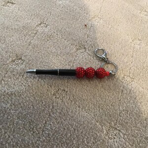 May include: A black pen with a silver tip and a decorative red beaded accent. The pen has a silver keyring attached to the end. The pen is lying on a beige carpet.