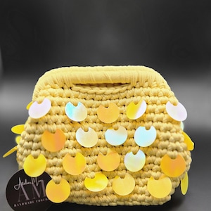 Puede incluir: Un bolso amarillo tejido a ganchillo con lentejuelas iridiscentes. El bolso tiene forma rectangular con asa y está decorado con múltiples lentejuelas amarillas e iridiscentes. La etiqueta dice "Atelier AM Handmade Crochet".