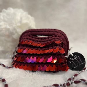 May include: A burgundy crochet handbag with a row of red sequins. The bag has a silver chain strap and a tag that reads "Atelier Mimi Handmade Crochet". The bag is on a white, fluffy surface.