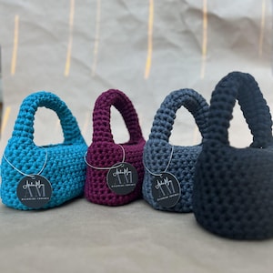 May include: Four small, handmade crochet bags in shades of teal, burgundy, gray, and black. Each bag has a rounded handle and a tag with the initials "AM". The bags are displayed on a neutral surface.