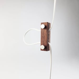 May include: A wooden cable organizer with a dark brown finish, featuring two silver-colored fasteners. White cables are neatly arranged through the organizer, creating a clean and minimalist aesthetic. The design is simple and functional.
