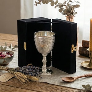 Könnte beinhalten: Ein silberner Kelch mit aufwendigen Blumen- und Herzmustern, präsentiert in einer offenen schwarzen Samtbox. Der Kelch steht auf einem Holztisch mit Lavendel, Weizen und einem Holzlöffel.