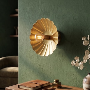 May include: A gold-toned wall sconce featuring a scalloped, flower-like metal shade with a central exposed filament light bulb. The fixture is mounted on a textured green wall, providing a warm glow from the centre of the circular metal petals.