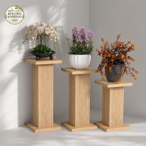 May include: Three light-colored wooden plant stands with vertical fluted columns and square bases. Each stand holds a potted plant: white orchids in a dark bowl, purple flowers in a white fluted pot, and orange foliage in a dark textured pot.