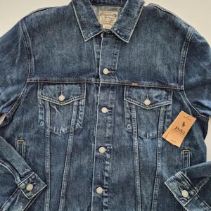 Puede incluir: Chaqueta vaquera azul medio con cierre de botones, dos bolsillos en el pecho y puños abotonados. La chaqueta tiene un cuello clásico y una pequeña etiqueta con el logotipo de la marca. Estilo clásico.