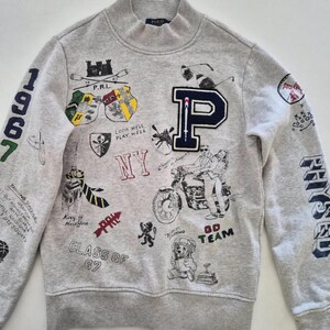 Puede incluir: Una sudadera gris claro de cuello alto con un gran parche de la letra P en azul marino y varios gráficos impresos que incluyen escudos, una motocicleta, un oso y los textos 1967, NY, Class of 67, Go Team, King of New York y Look Well Play Well.