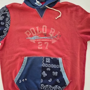 May include: Red hoodie with a navy blue hood and pocket. The hoodie features the text "POLO" in a distressed font, along with the number "27". The pocket and one sleeve have a paisley pattern.