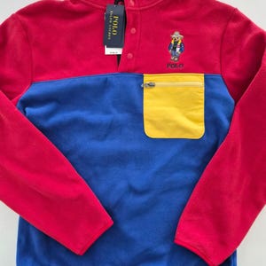 Puede incluir: Un jersey polar con bloques de color rojo, azul y amarillo. La sección superior es roja, la del medio es azul y un bolsillo amarillo está en la parte inferior delantera. Cuenta con un cuello alto con botones y el logo Polo bear.