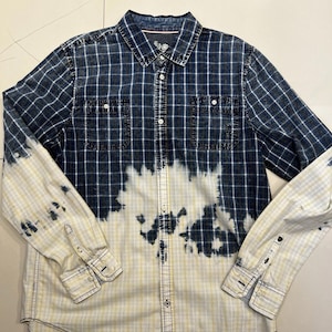 Może przedstawiać: Koszula z długim rękawem w niebiesko-białą kratę. Dolna połowa koszuli i rękawy są wybielone, tworząc unikalny dwutonowy efekt. Posiada dwie kieszenie na piersi i mankiety zapinane na guziki.