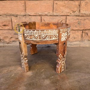 May include: A handcrafted wooden plant stand with a round top and four legs. The stand features intricate carved designs and white accents. The wood has a warm, natural finish, and the stand is designed to hold a potted plant.