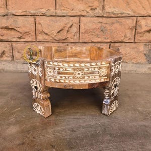 May include: A handcrafted wooden decorative bowl with carved floral and geometric patterns. The bowl has a light brown finish with white accents and is supported by four carved legs. The bowl is designed to hold small items.