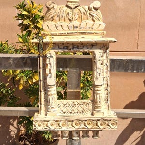 Puede incluir: Marco de ventana de madera tallada de color crema, con detalles florales y geométricos. El marco presenta una parte superior e inferior decorativas, con una abertura central. El texto "Pawan Art And Craft" es visible.