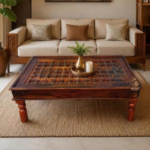 May include: A square wooden coffee table featuring a dark brown finish, a grid-patterned top with embedded metal studs, and four turned legs. The table surface displays a weathered texture and is adorned with a small brass vase and a white candle.