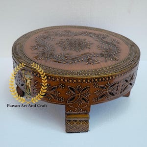 May include: A round, brown wooden decorative stand with intricate floral carvings. The stand has a detailed border and is supported by short, square legs. The text "Pawan Art And Craft" is visible on the side.