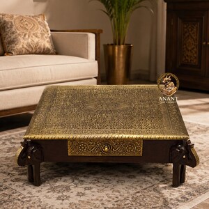 May include: A square coffee table featuring a dark wood frame and a brass top with intricate floral engravings. The table includes a central drawer with a matching brass front and four legs carved into the shape of elephant heads and trunks.