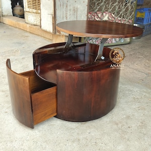 May include: A round dark brown wooden coffee table with a lift-top mechanism and a curved pull-out drawer. The table features a smooth polished finish and a gold-coloured circular logo with the text ANANT ART EXPORT printed on the side.
