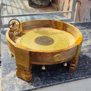 Op de afbeelding: Een rond houten dienblad met een natuurlijke afwerking, voorzien van een centrale goudkleurige metalen inleg met ingewikkelde reliëfpatronen. De buitenkanten zijn versierd met goudkleurige metalen platen en kleine hangende belletjes, op vier poten.