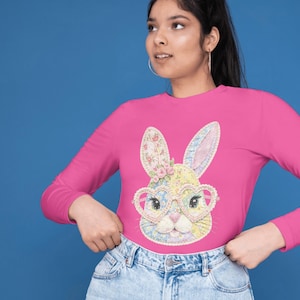 May include: A long-sleeved, hot pink shirt featuring a colorful bunny face with heart-shaped glasses. The bunny design incorporates floral and pastel patterns. The shirt is paired with blue denim jeans.