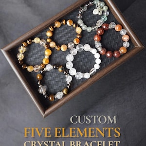 May include: Five beaded bracelets made of various natural crystals, including tiger eye, clear quartz, and reddish-brown stones, arranged in a dark wooden tray. The text reads: CUSTOM FIVE ELEMENTS CRYSTAL BRACELET PERSONALIZED BY BIRTH DETAILS.