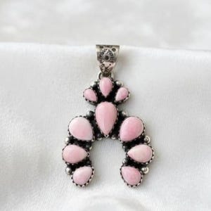 Könnte beinhalten: Ein silberfarbener Anhänger mit hufeisenförmigem Design, besetzt mit tropfenförmigen rosa Steinen. Der Anhänger hat eine dekorative Öse und ist vor einem weißen Hintergrund platziert, um die Details des Schmucks hervorzuheben.